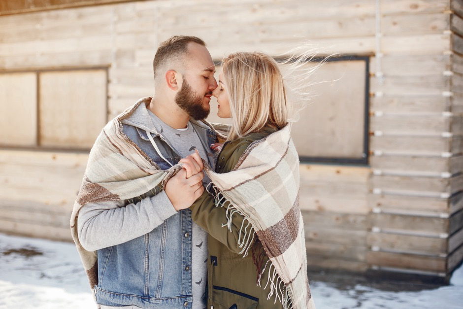 man and woman wearing a blanket embrace outside a snowy cabin