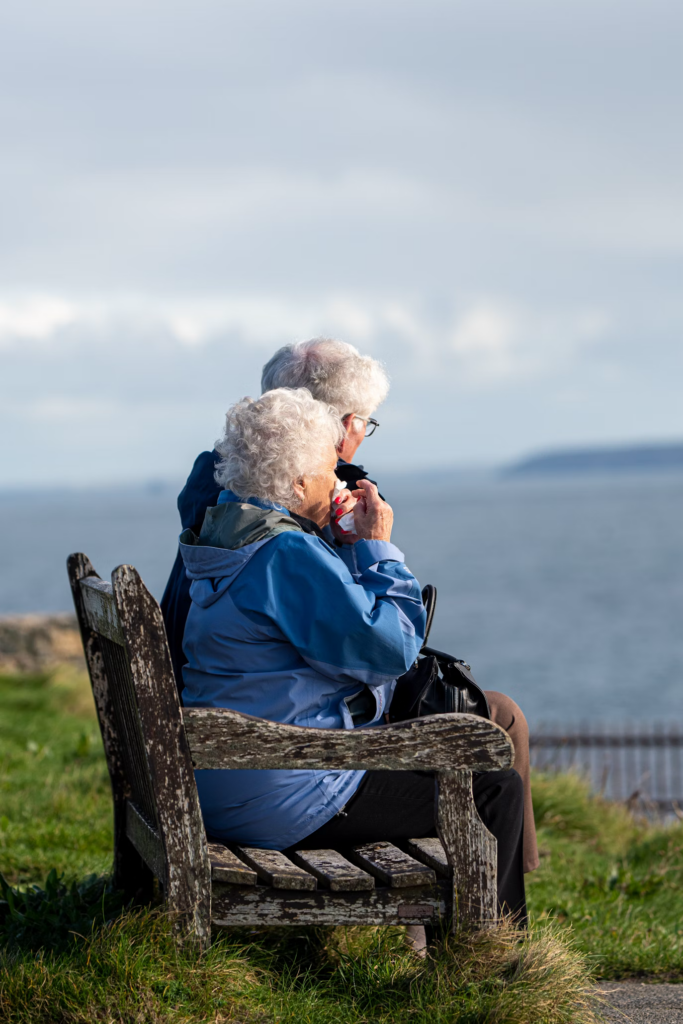 An elderly couple sitting outdoors enjoying nature, surrounded by greenery.