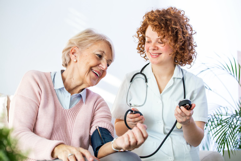 A young nurse with curly hair checks an elderly woman’s blood pressure.