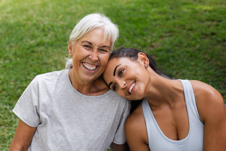 An older woman smiles as a younger woman lays her head on the older woman’s shoulder.